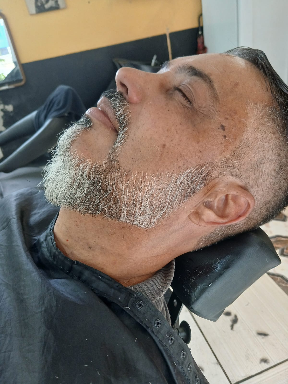cliente de barba grisalho reclinado na cadeira com barba aparada e cabelo penteado